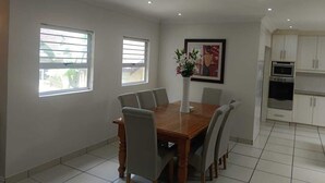 A dining area with a wooden table and six chairs, two windows with blinds, a framed picture on the wall, and a kitchen with white cabinets and appliances. - Pioneer Guest House (DURBAN)