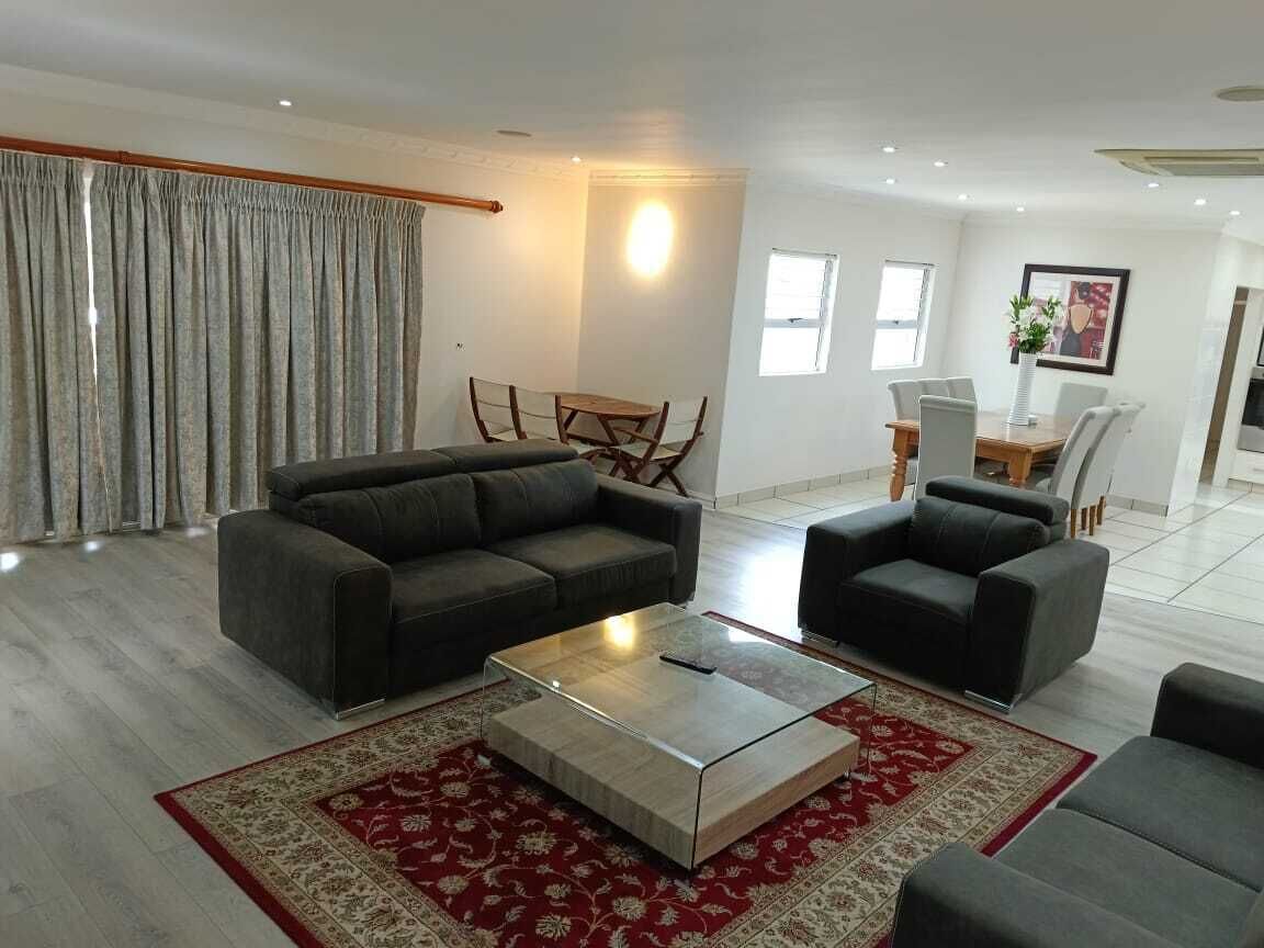 A modern living room with a grey sofa set, a glass coffee table, and a red patterned rug. There is a dining area with a wooden table and chairs, and a kitchen area in the background.