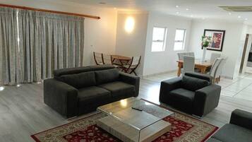 A modern living room with a grey sofa set, a glass coffee table, and a red patterned rug. There is a dining area with a wooden table and chairs, and a kitchen area in the background.