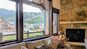 Ein Wohnzimmer mit großem Fenster, Kamin aus Stein und Blick auf eine Wohngegend mit Häusern und Bergen.