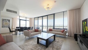 A modern living room with two sofas, a coffee table, and a TV. There is a dining area with chairs and a table, and large windows offering a view. - Silk Valley-4B with Private Beach Access (Al Hamriya)