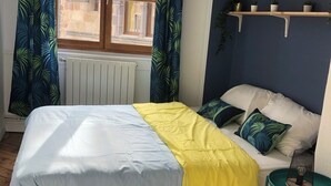A bedroom with a bed, a window with curtains, a radiator, a shelf with plants, and a small table with a lamp. - Welcome to this large, elegant apartment. (Brioude)