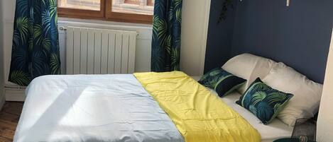 Ein Schlafzimmer mit einem Bett, einem Fenster mit Vorhängen, einer Heizung, einem Regal mit Pflanzen und einem kleinen Tisch mit einer Lampe.