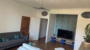 A living room with a sofa, a television on a wooden stand, a ceiling fan, and a door leading to another room. - Welcome to this large, elegant apartment. (Brioude)