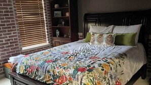 A bedroom with a large bed, a wooden dresser, a brick wall, and a window with blinds.