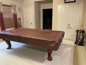 Une table de billard dans une pièce dont le sol est recouvert d’une moquette beige, où l’on trouve des portes blanches et une fenêtre avec des stores.