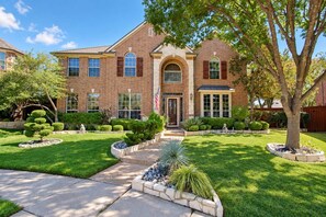 A two-story brick house with a front entrance, a flag, and a well-maintained lawn with various shrubs and a tree.