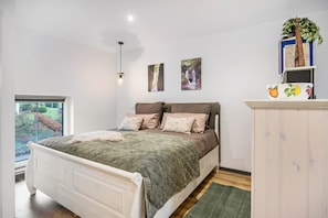 Un dormitorio con una cama, una ventana, un estante en la pared con libros y una planta, y un cuadro.