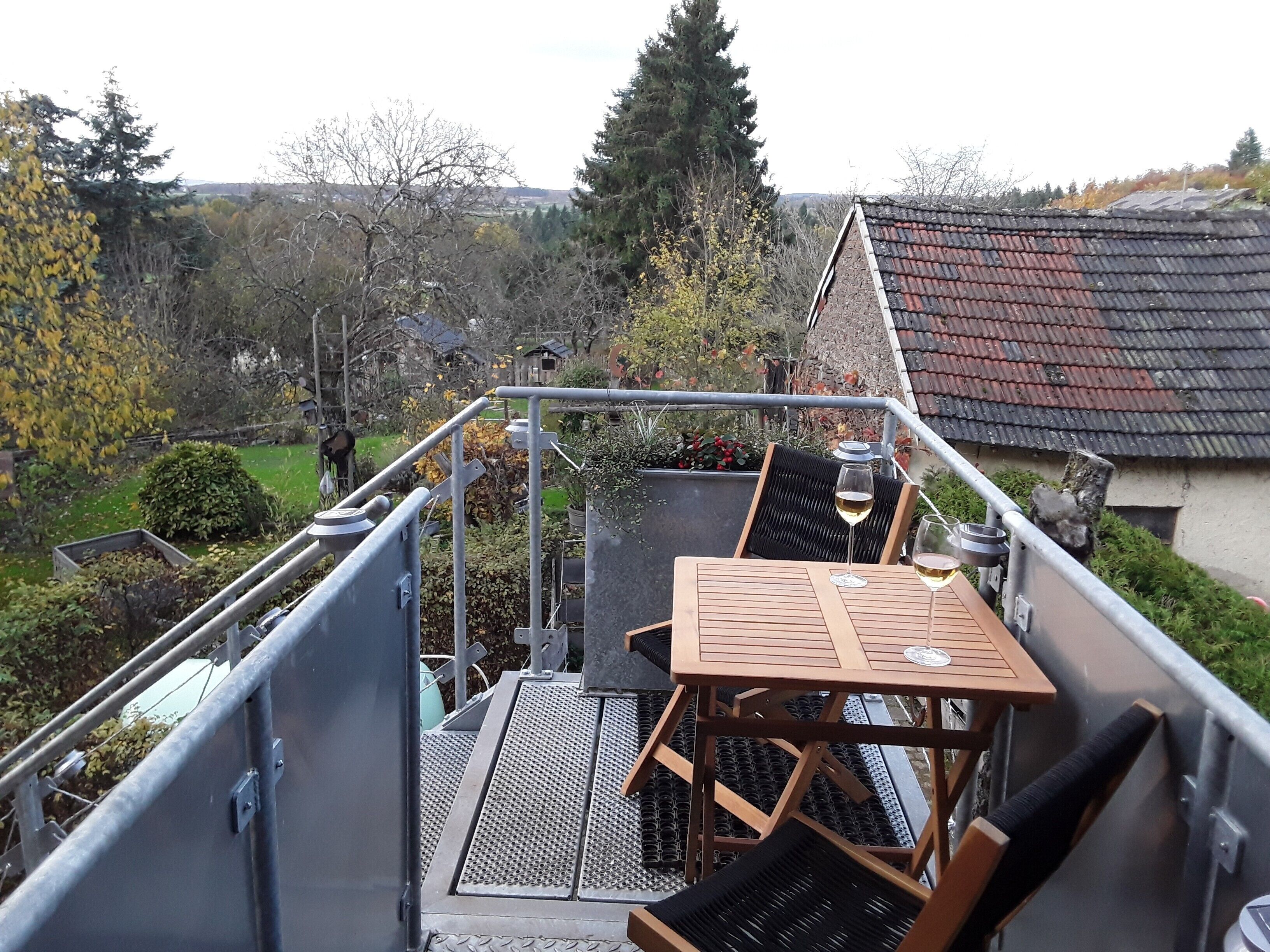 Una escalera metálica conduce a una terraza en la azotea con una mesa y sillas, rodeada de vegetación y casas al fondo.