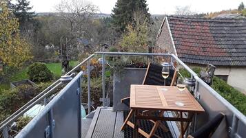 Una escalera metálica conduce a una terraza en la azotea con una mesa y sillas, rodeada de vegetación y casas al fondo.