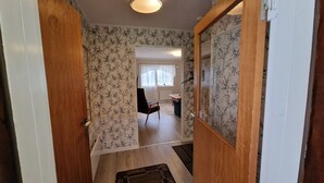 Un couloir avec un papier peint à motifs, une porte en bois et une vue sur une pièce où se trouvent un bureau et une chaise.