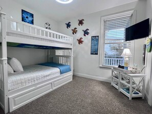 A bunk bed room with a desk and bookshelf, a window with blinds, and wall decorations including a life preserver and sea turtle.