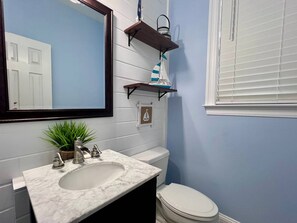 A bathroom with a white sink, a mirror, a toilet, and a window with blinds. - Welcome to Sea Shed – Private Pool, Fire Pit, Pet Friendly (Cape San Blas)