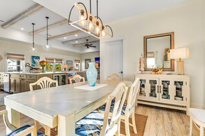 A spacious dining area with a large table and chairs, a modern chandelier, and a kitchen in the background.