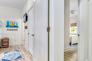 A hallway with a wooden floor, a blue patterned rug, a painting of sailboats, and a bed in the adjacent room.