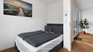 A bedroom with a bed, a large landscape painting, a potted plant, and a view through a glass door.