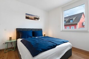 Un dormitorio con cama, mesita de noche, un cuadro en la pared y ventana con vistas a edificios.