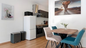 A compact kitchen with a microwave, oven, and a small refrigerator. There is a dining area with a wooden table and chairs, and a painting of an elephant on the wall.