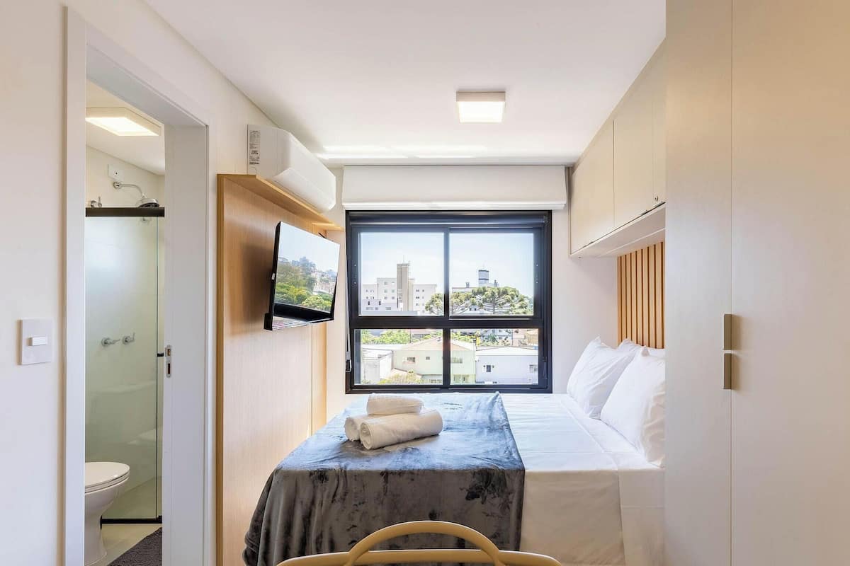 Ein schmales Hotelzimmer mit Bett, einem an der Wand montierten Fernseher, einem Fenster mit Blick auf die Stadt und einer Dusche.