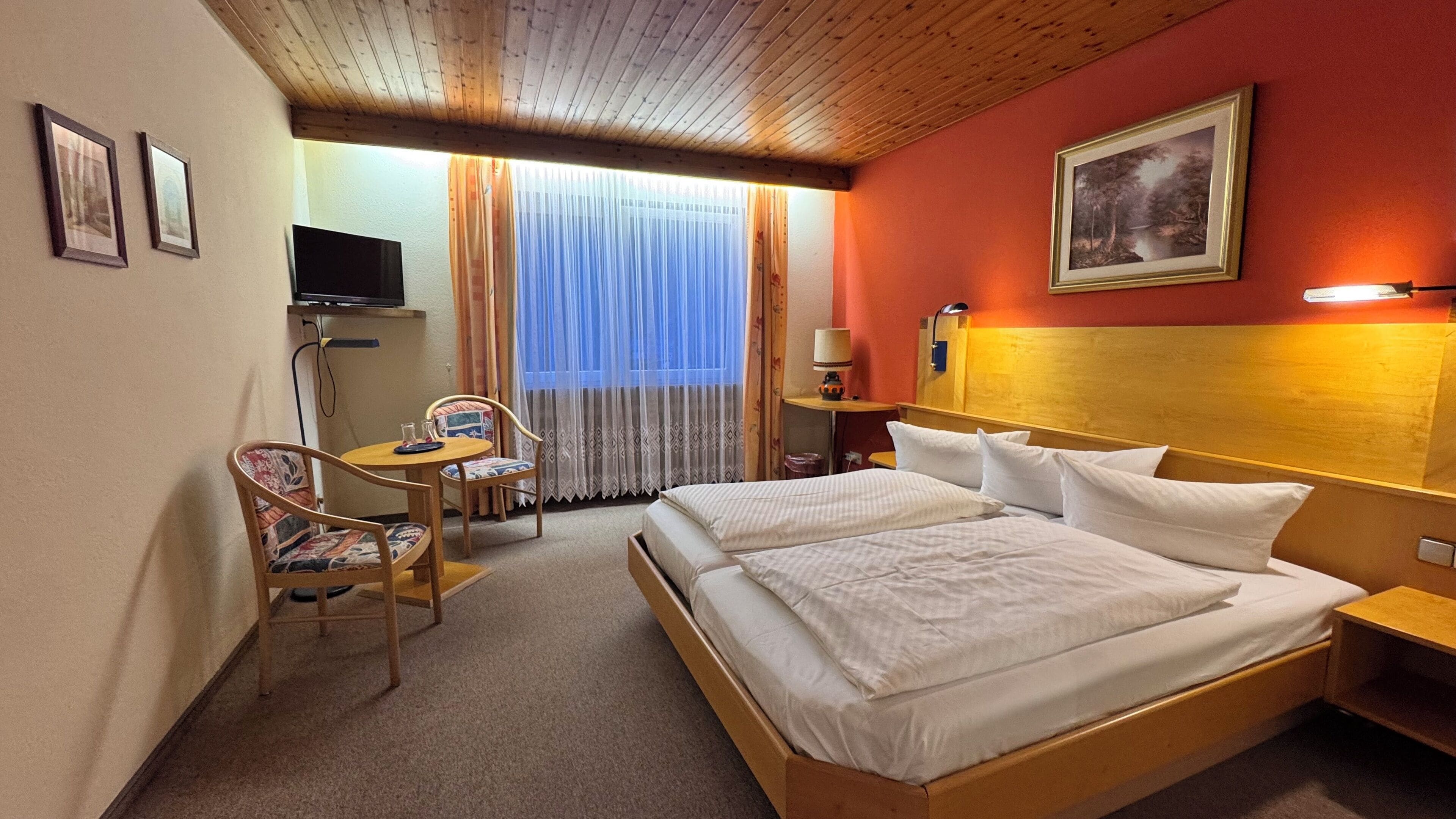 Ein Hotelzimmer mit einem hölzernen Bett, einem Schreibtisch mit Stuhl, einem Fernseher und einem Fenster mit Vorhängen.