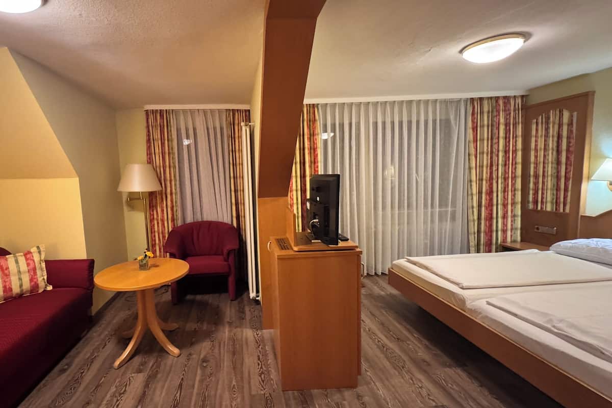 Ein Hotelzimmer mit Holzboden, einem Bett, einem roten Sofa, einem kleinen Tisch mit einer Lampe und einem Fernseher auf einem Holztisch.