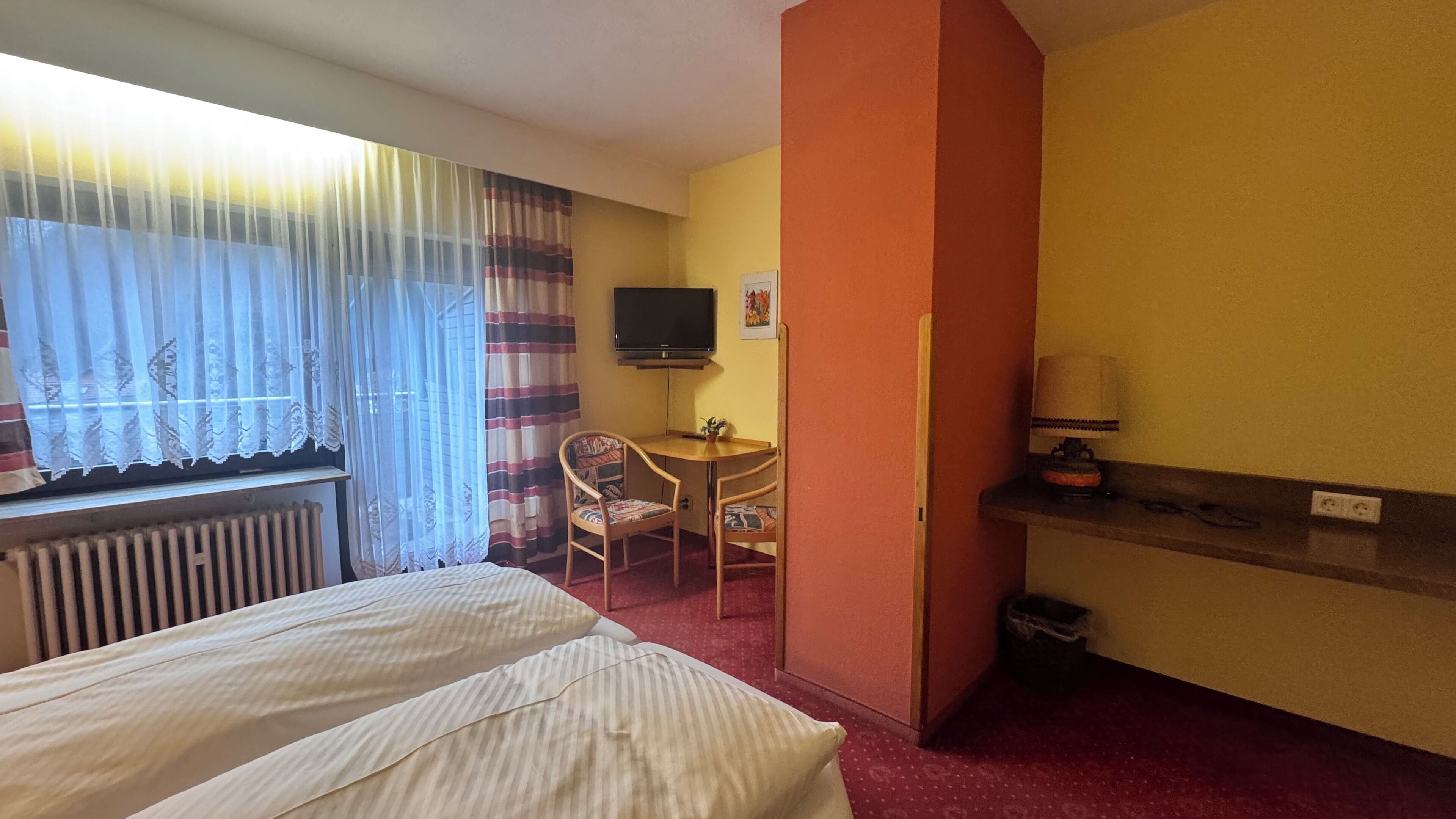 A hotel room with a bed, a desk with a chair, a television, and a radiator.