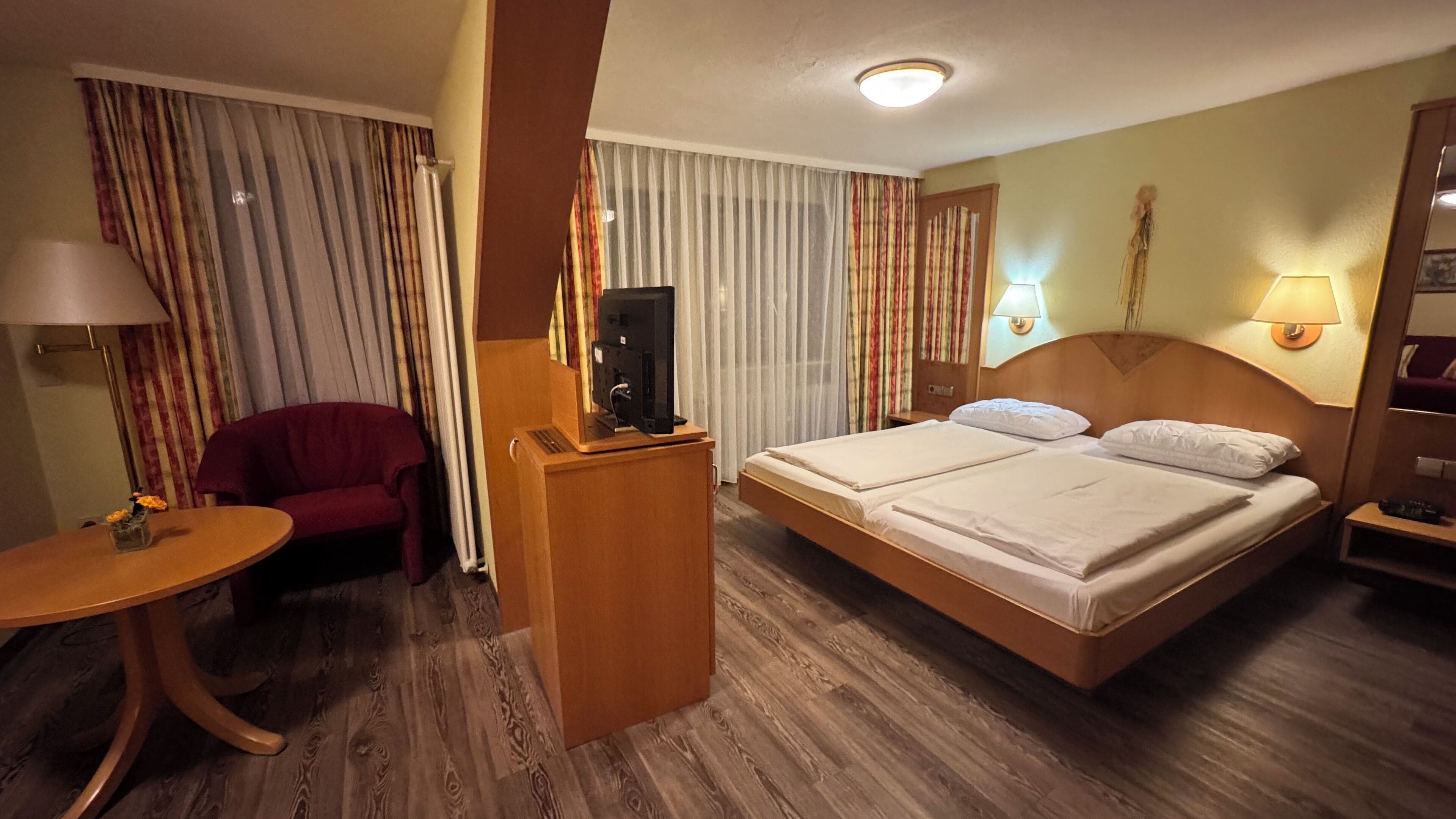 A hotel room with a double bed, a desk with a television, a red armchair, and a small table with a vase of flowers.