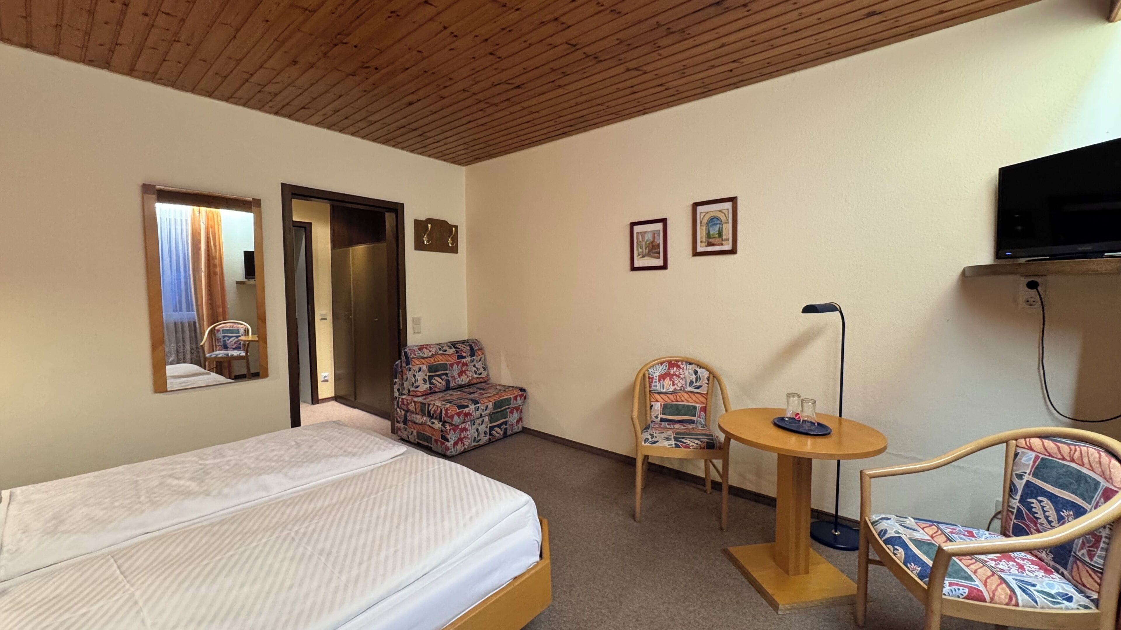 Habitación de hotel con una cama, una silla, una mesita, un televisor y una puerta que da a otra habitación.