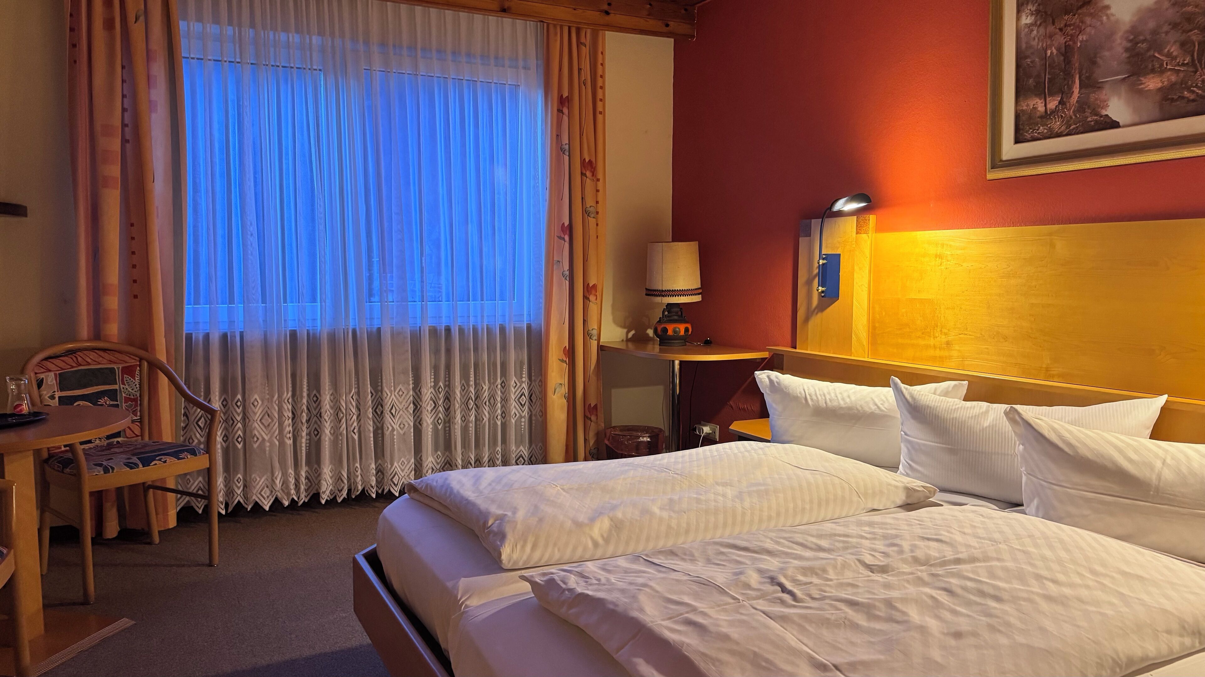 A hotel room with a bed, a desk with a chair, a lamp, and a painting on the wall.