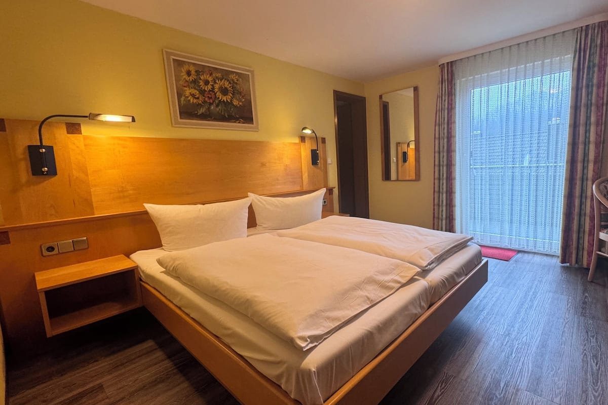 Ein Hotelzimmer mit einem großen Bett, Nachttischlampen, einem kleinen Tisch, einem Sessel, einem Bild an der Wand und einem Fenster mit Vorhängen.