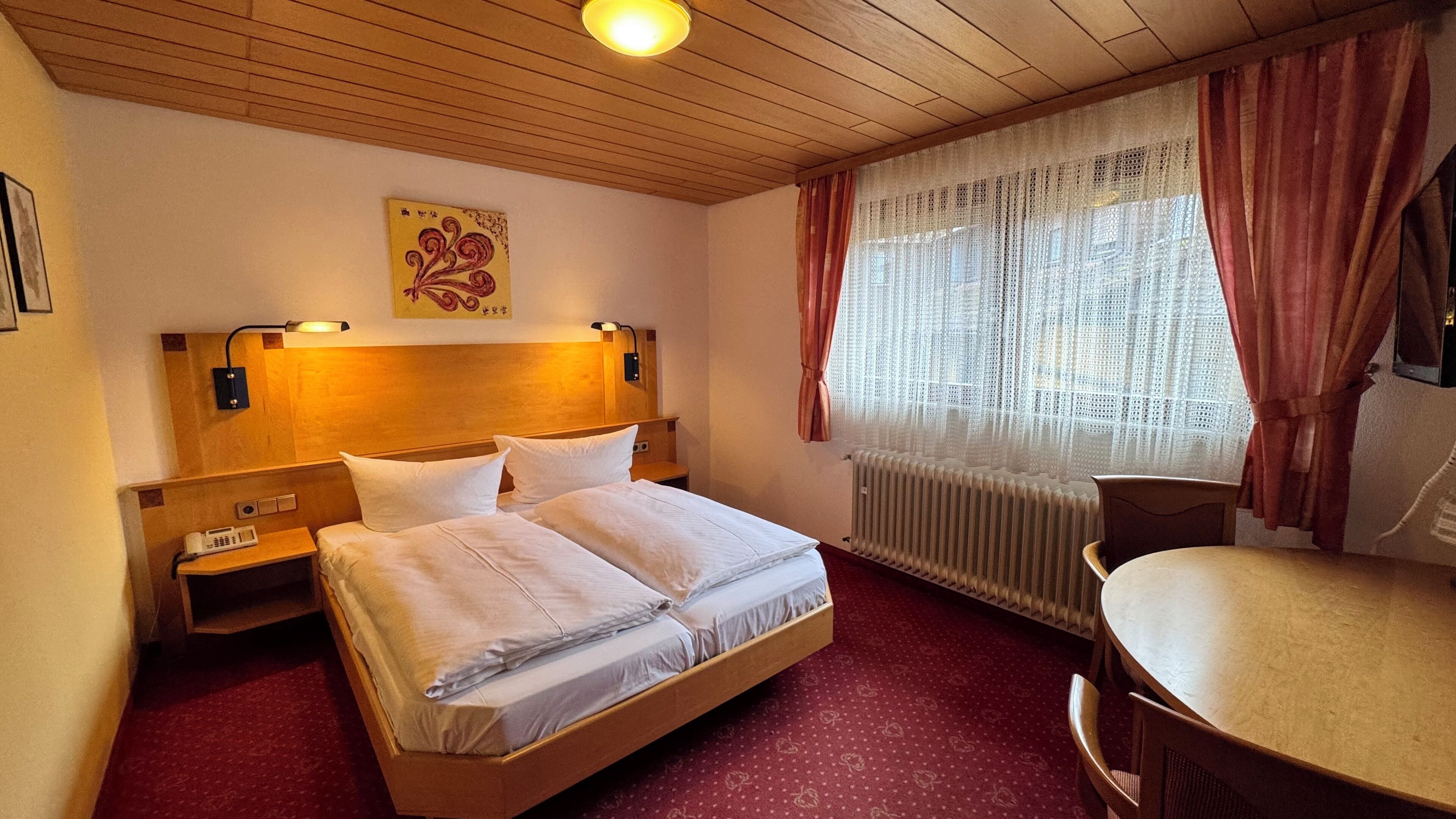 A hotel room with a wooden headboard, a bed with white linens, a bedside table with a phone, a radiator, a window with sheer curtains, and a small round table.