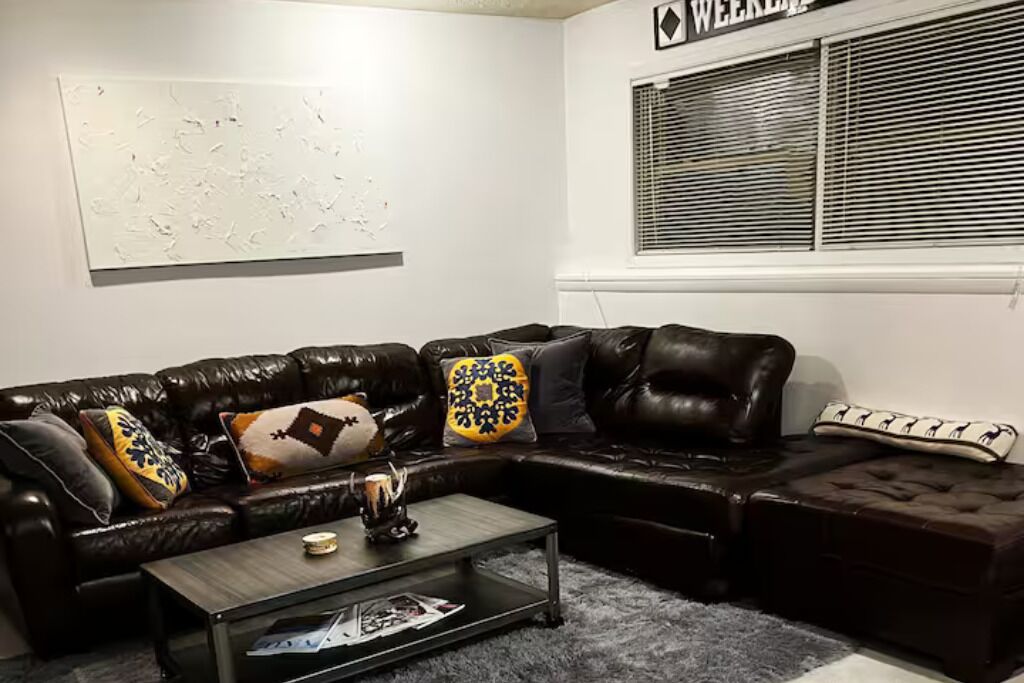 A living room with a dark brown leather sectional sofa, a glass coffee table, and a window with blinds.