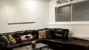 Una sala de estar con un sofá seccional de cuero marrón oscuro, una mesa de centro de vidrio y una ventana con persianas.