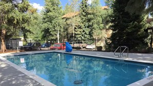 Una piscina con una escalera metálica, rodeada de árboles y coches estacionados.