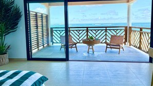 Una habitación con un gran ventanal corredera que da a un balcón con vista al océano. Hay dos sillas de mimbre y una mesita en el balcón.