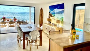 Un comedor moderno con una mesa grande y sillas, una tabla de surf de madera, y vista al océano a través de amplios ventanales.