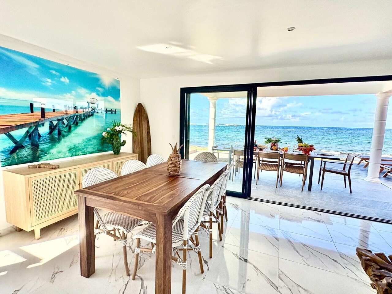 A dining area with a wooden table and wicker chairs, overlooking a beach with a pier.