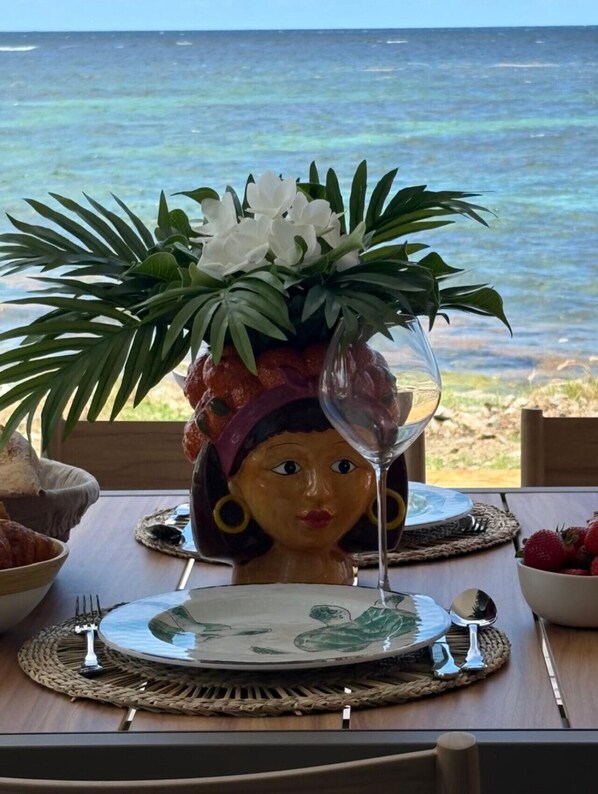 A table set with a decorative ceramic figure, a glass, and a plate with a green design, overlooking a beach.