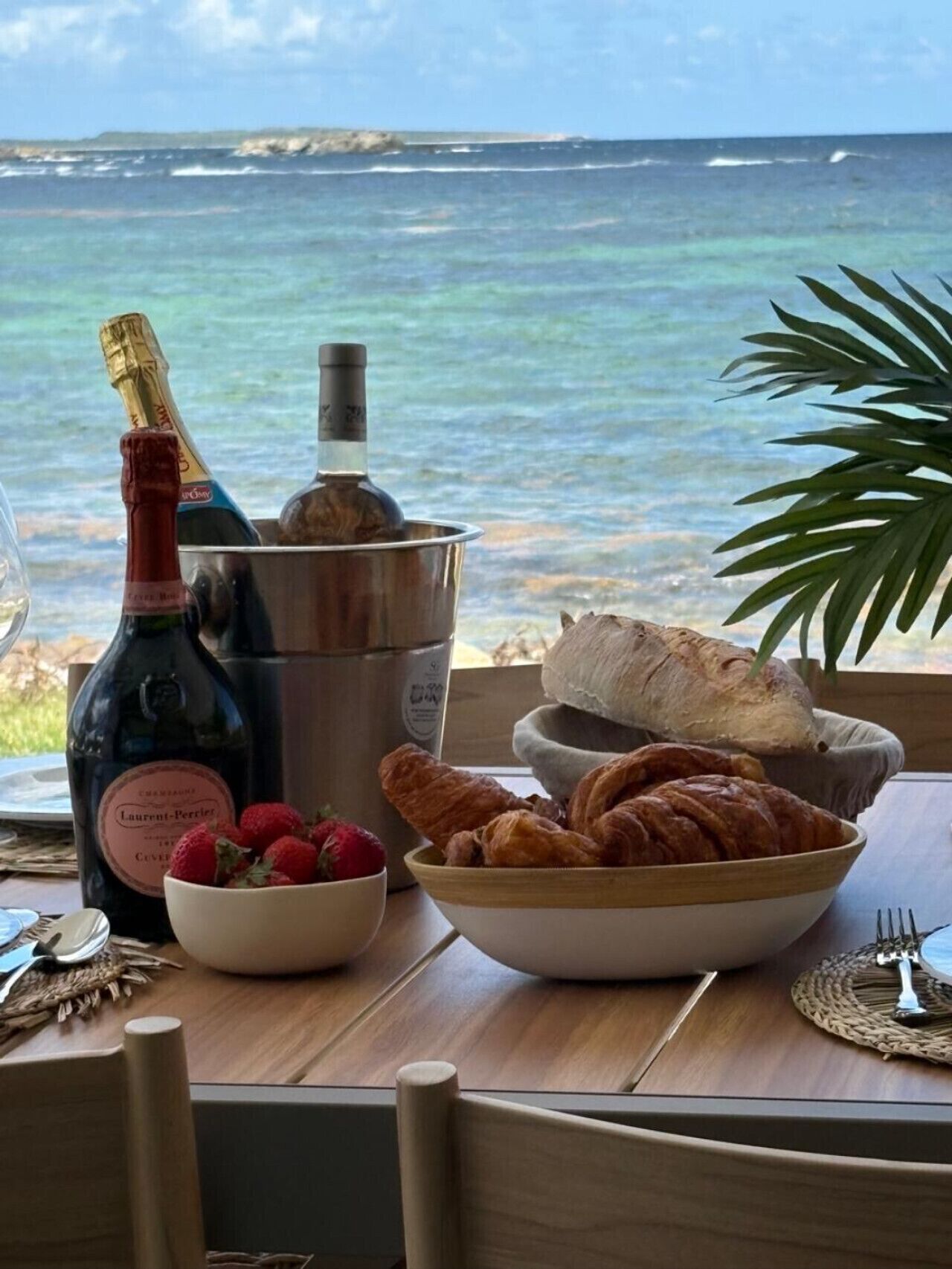 Ein Tisch mit einer Weinflasche, einem Eimer, einer Schale Erdbeeren und Croissants vor einem Fenster mit Meerblick.