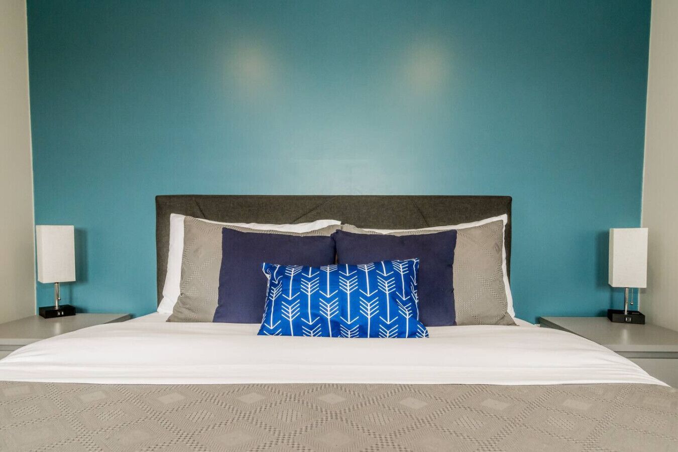 Una cama con cabecera gris, ropa de cama blanca y una almohada con estampado azul. Hay dos mesitas de noche con lámparas a ambos lados de la cama.
