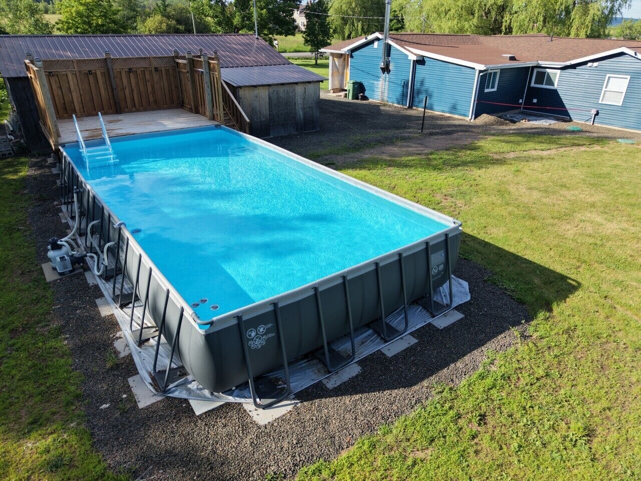 An above-ground swimming pool with a ladder, surrounded by a grassy yard and wooden fencing.