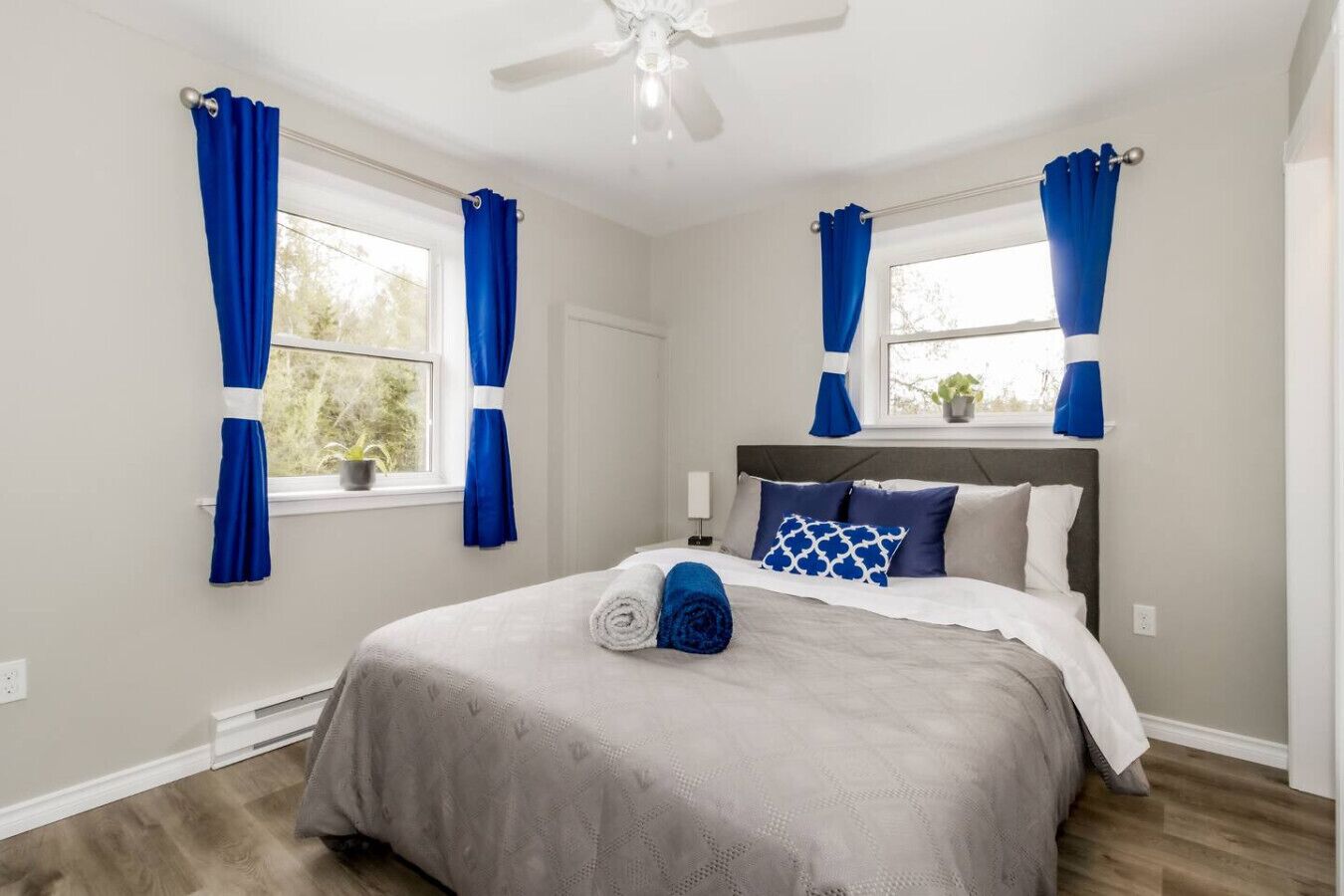 A bedroom with a bed, two windows with curtains, a ceiling fan, and a nightstand.