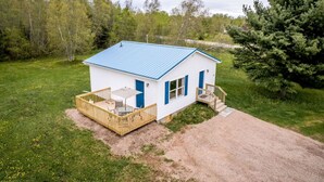 A small, single-story cabin with a blue roof, a deck, and a dirt driveway.