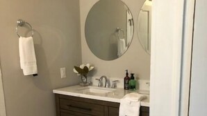 A bathroom with a marble floor, a double sink vanity, and a round mirror. - Jacklin Cottage at Generals' Quarters (Corinth)