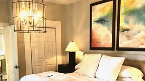 A bedroom with a large bed, a chandelier, a nightstand with a lamp, and two abstract paintings on the wall. - Jacklin Cottage at Generals' Quarters (Corinth)