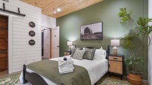 A neatly arranged bedroom with a bed, bedside tables, a wall clock, a painting, and a potted plant.