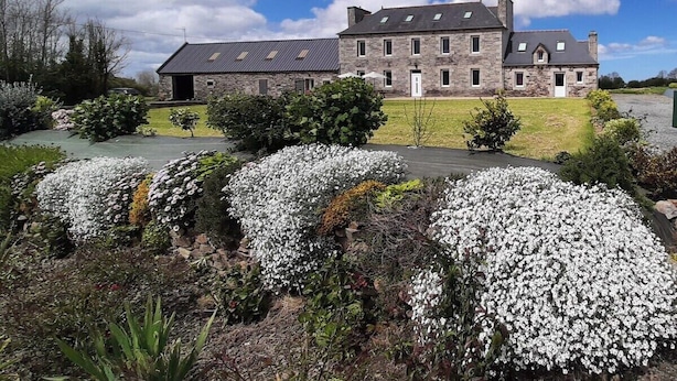 Un grand bâtiment en pierre, doté de nombreuses fenêtres et d’une entrée centrale, entouré de jardins bien entretenus et de plantes à fleurs blanches.