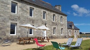 Un bâtiment en pierre doté d’un patio comprenant une table en bois et deux parasols, ainsi que deux chaises longues.