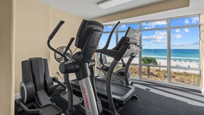 Une salle de sport équipée de tapis de course et de vélos d’exercice, avec de grandes fenêtres donnant sur la plage.