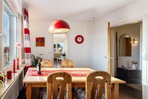 A dining area with a wooden table, chairs, a red pendant light, a clock, and a view through a window.